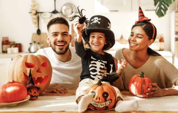 Cheerful multi ethnic family parents with son smiling while creating jack o lantern from pumpkin during Halloween celebration in kitchen at home