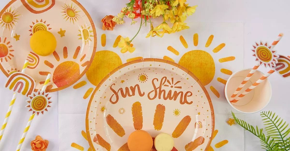 A bright, cheerful arrangement of paper plates, cups, napkins, and straws decorated with sun motifs, rainbows, and warm orange and yellow hue