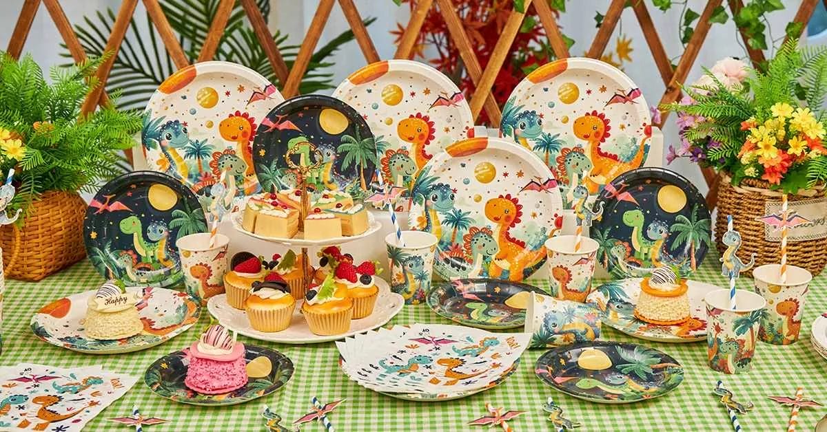 A vibrant party setup with plates, cups, napkins, and toppers featuring colorful cartoon dinosaurs, palm trees, and a prehistoric jungle back